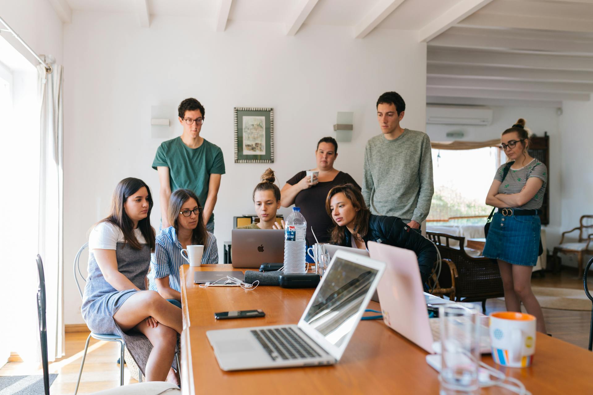 A group of people gathered around a table with laptops and notebooks, while engaging in a discussion in a bright room.