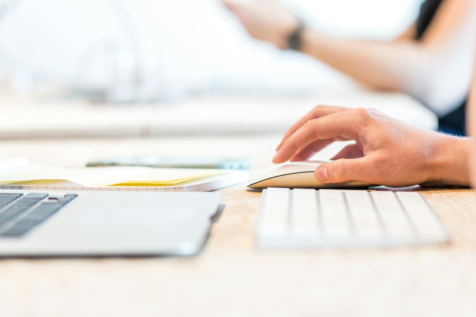 A person using a wireless mouse next to a keyboard and laptop on a desk.
