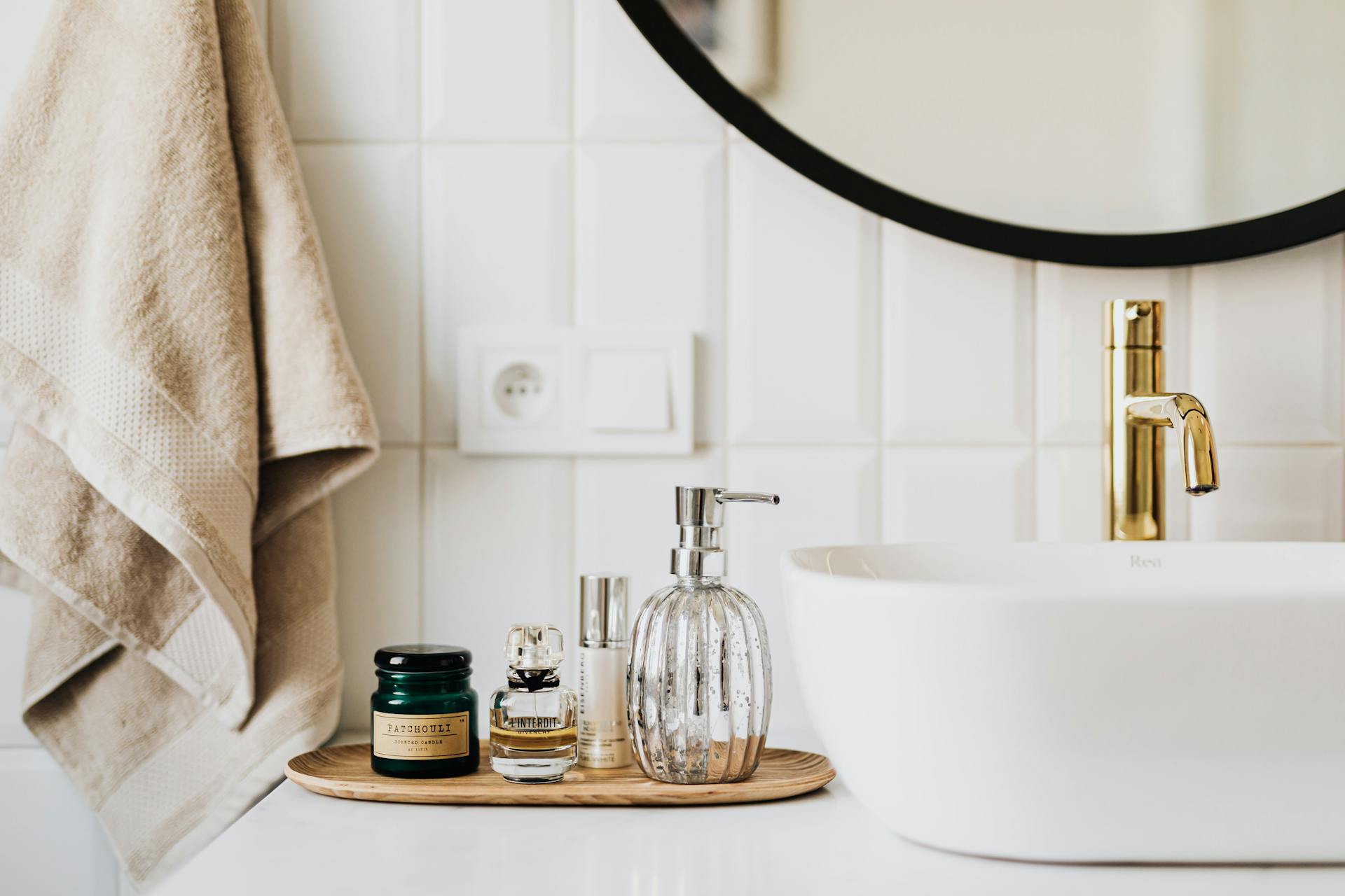 Bathroom counter with a round mirror, beige towel, and a tray holding skincare jars and a soap dispenser near a white sink with a gold faucet.