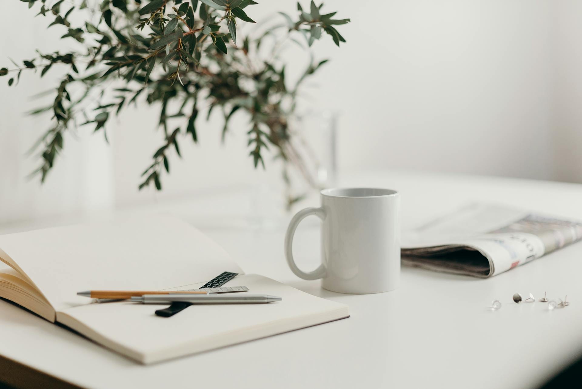 A white mug, open notebook with pens, a newspaper, and a vase with greenery sit on a white table.