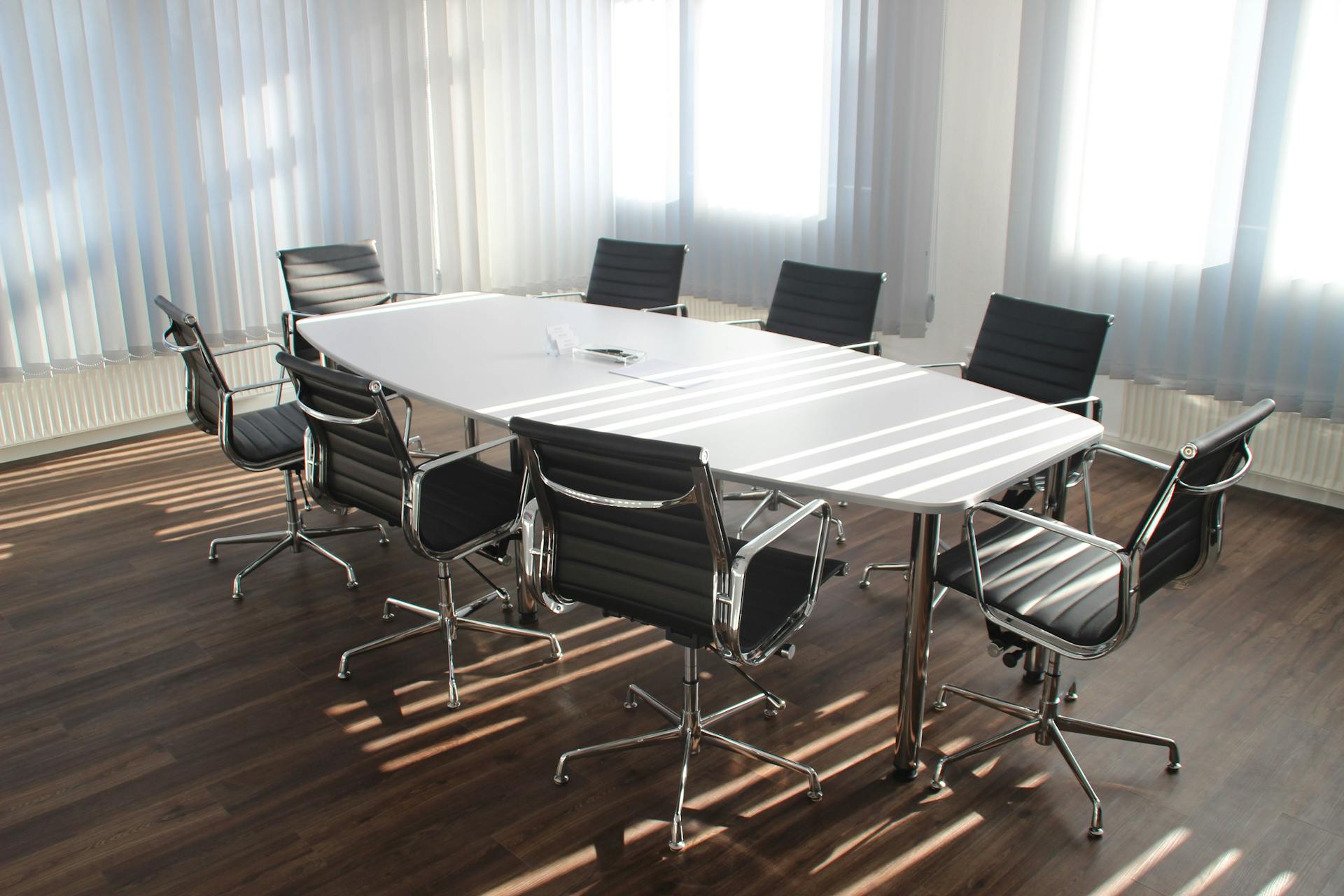 A long white conference table surrounded by eight black chairs in a meeting room with wooden flooring and vertical blinds on windows.