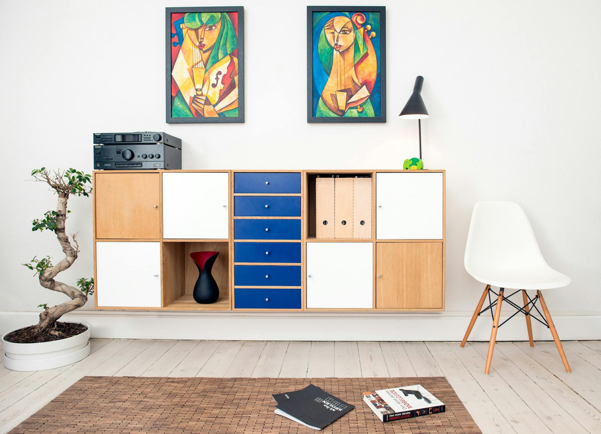 Modern living room with a wooden cabinet featuring blue and white doors, a bonsai, a white chair, and colorful artwork. A stereo sits on the cabinet. Books lie on a woven mat.