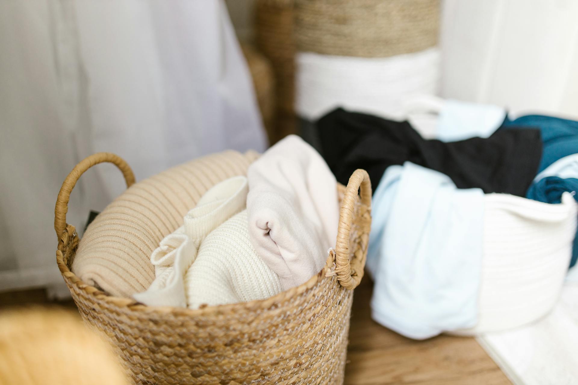 Two woven baskets filled with neatly folded clothes, including neutral and dark-colored garments, placed on a wooden floor.