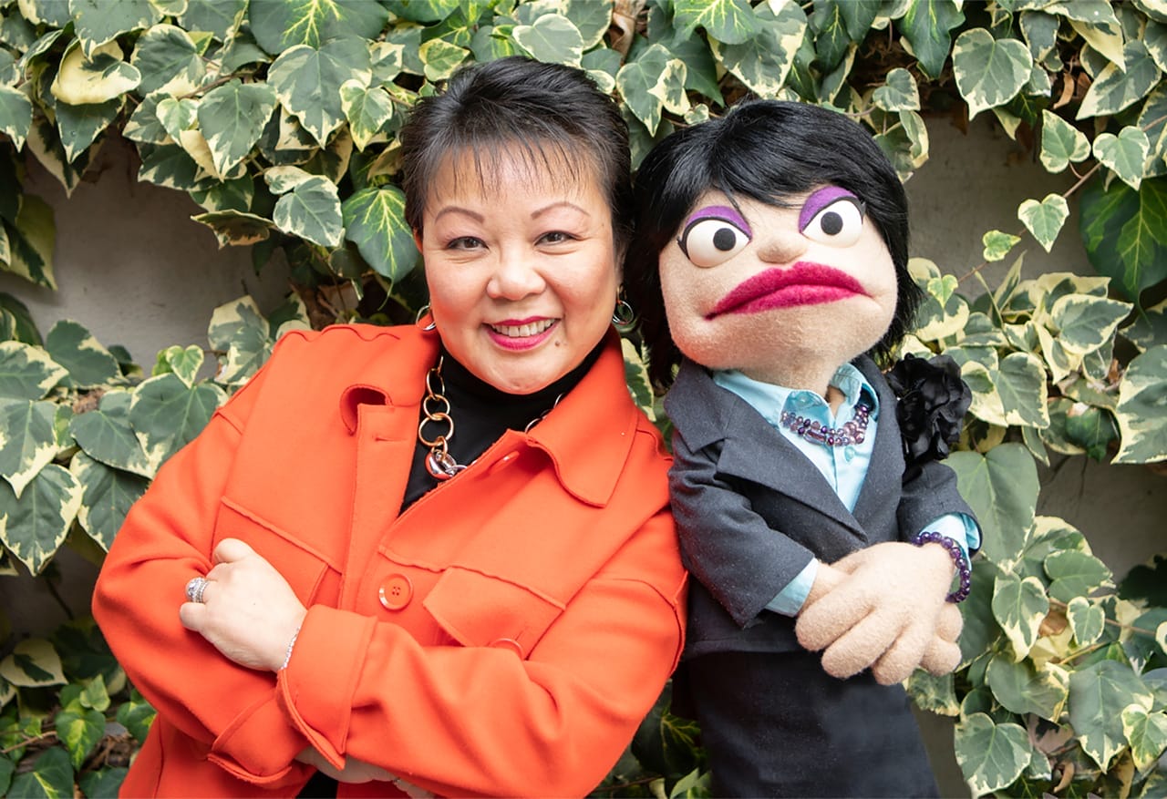 Linda Chu is in an orange coat and stands smiling with a puppet in a suit in front of a leafy background. Both have their arms crossed.