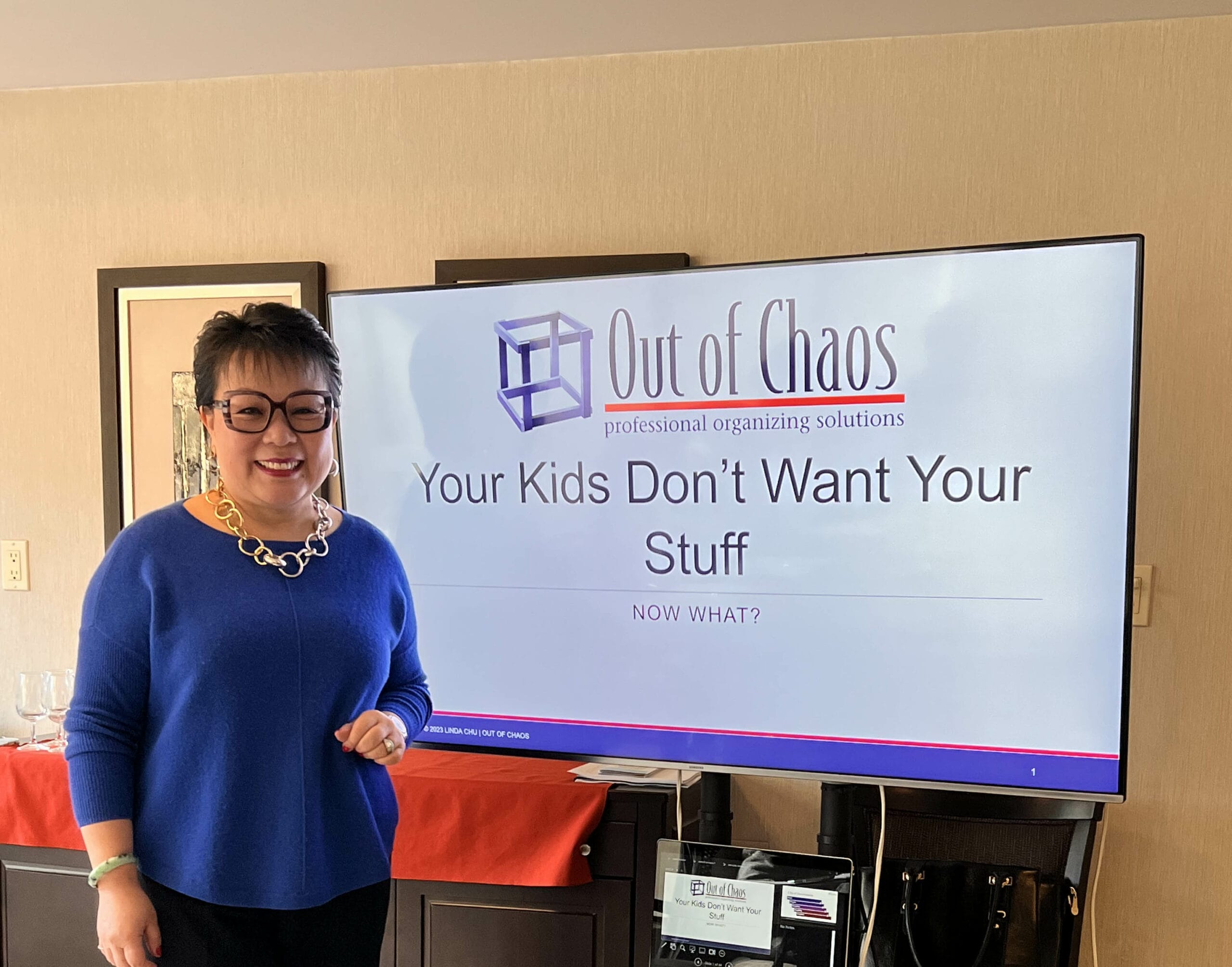 Linda Chu, wearing glasses and a blue sweater with a statement necklace, standing next to a presentation screen displaying the title 'Out of Chaos: Your Kids Don’t Want Your Stuff – Now What?' in a professional setting.