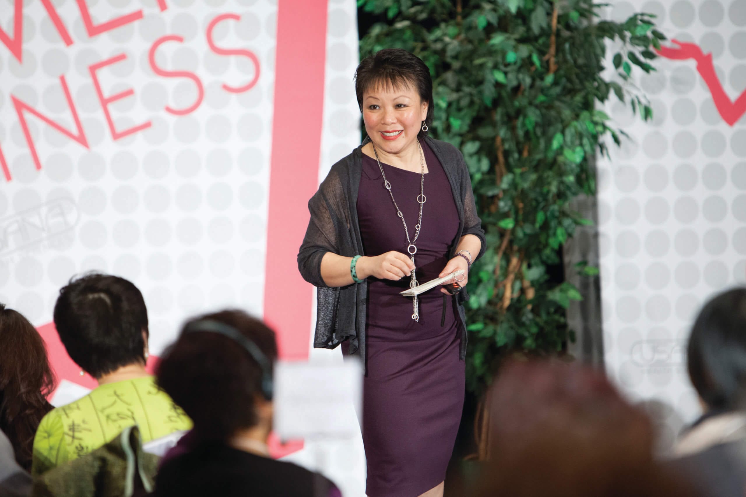 Linda Chu is in a purple dress and speaks to an audience at a business event, standing near a plant and a sign with partial text "WOMEN IN BUSINESS"