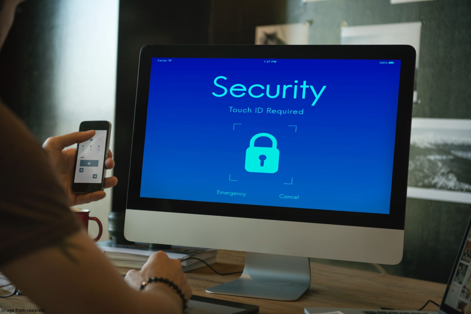 computer on desk showing screen with the word Security and a padlock and a person looking at a cell phone and the screen
