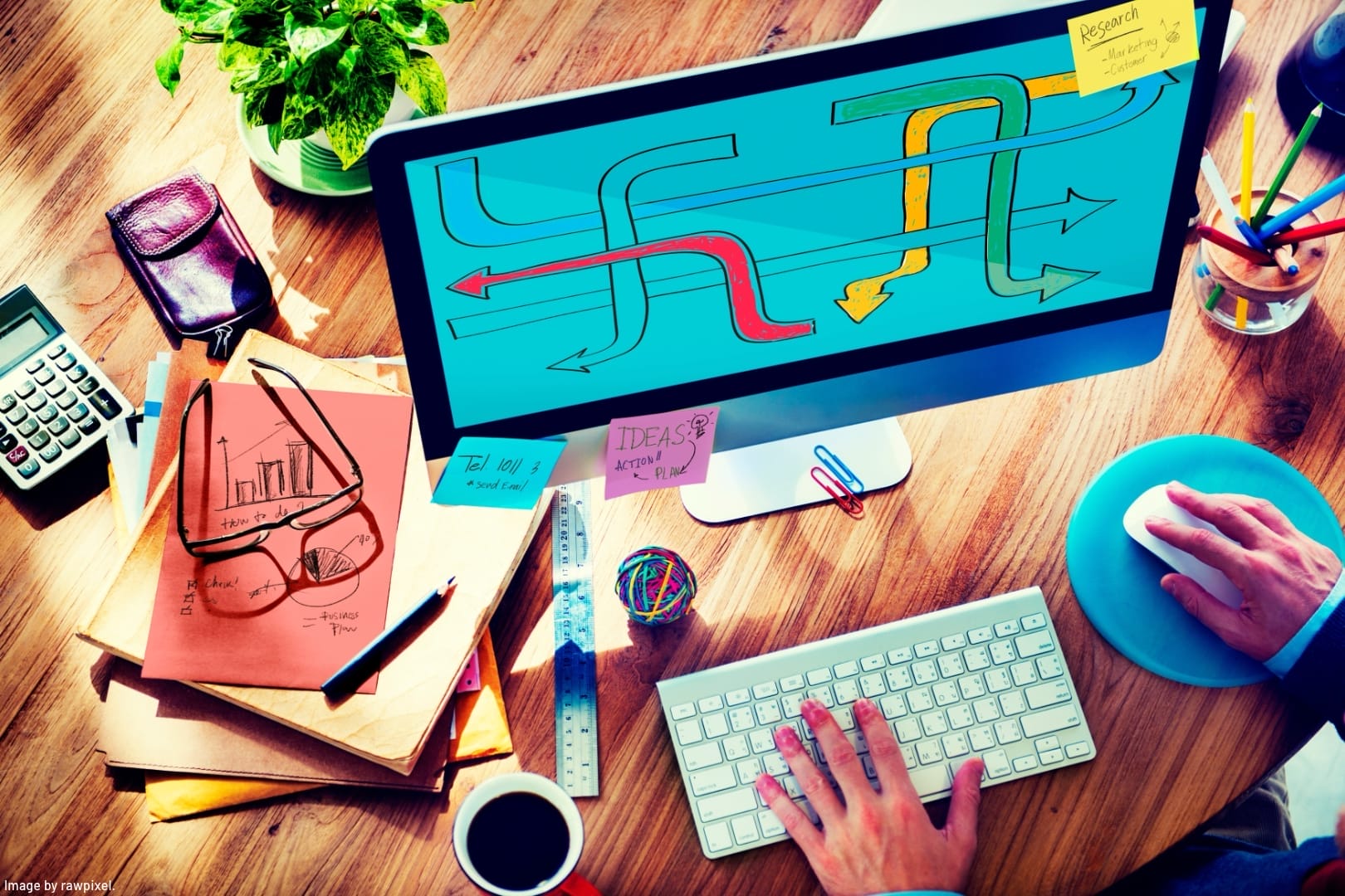 person in front of computer with one hand on keyboard other hand on mouse. desk piled with papers representing how to multitask successfully